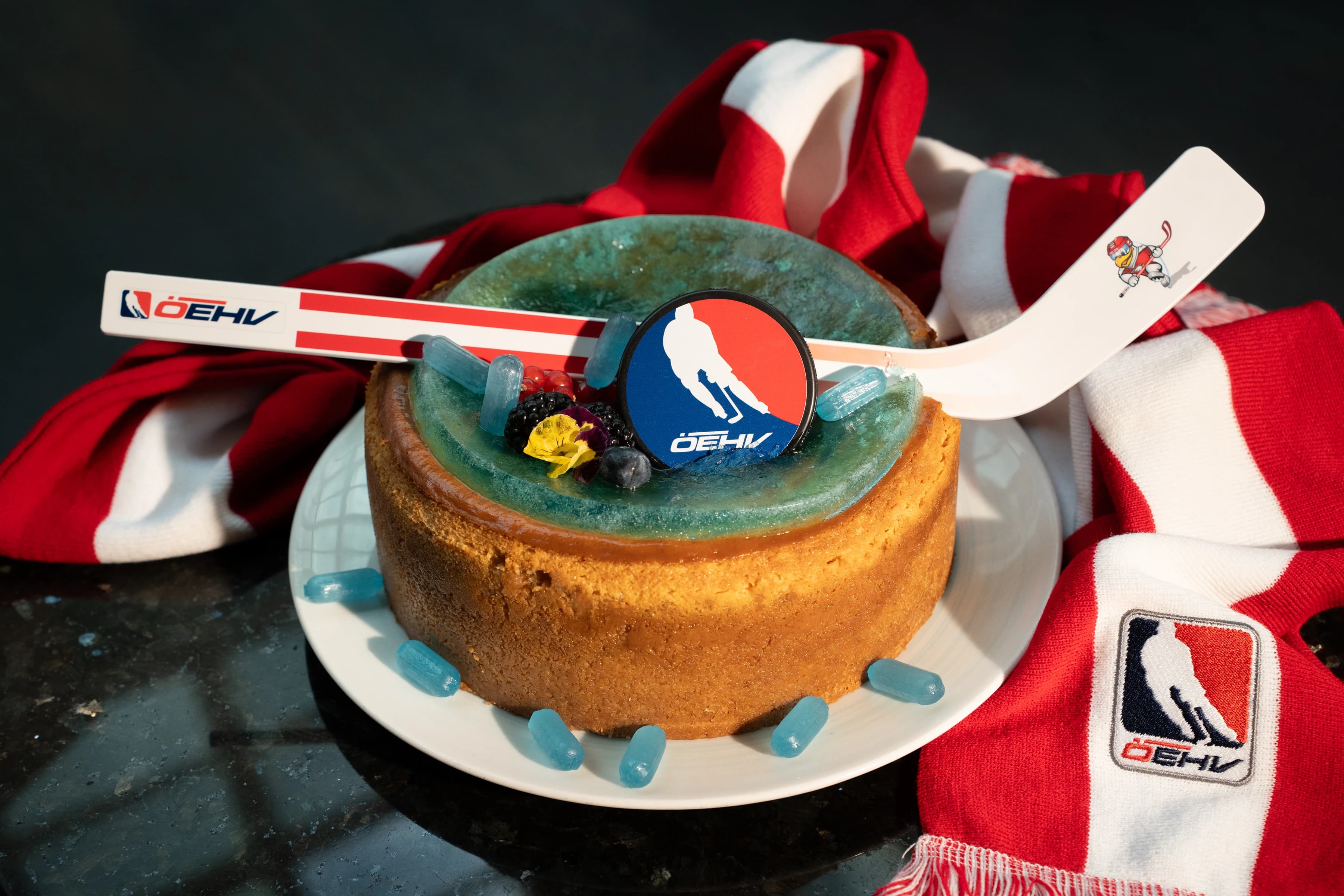Cheesecake mit ÖEHV-Branding: Ein runder Cheesecake mit goldbraunem Boden und blaugrünem Spiegelglasur-Topping, dekoriert mit Beeren, essbaren Blüten und blauen Eiswürfel-förmigen Zuckerdeko-Elementen. Auf dem Kuchen liegt ein Eishockeypuck mit ÖEHV-Logo, im Hintergrund ein Mini-Eishockeyschläger mit ÖEHV-Branding. Der Kuchen steht auf einem weißen Teller, umgeben von einem rot-weißen ÖEHV-Fanschal.